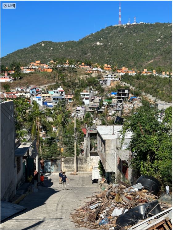 Looking down steep hill, damaged apartment buildings in background. 2 people walking up steep road.